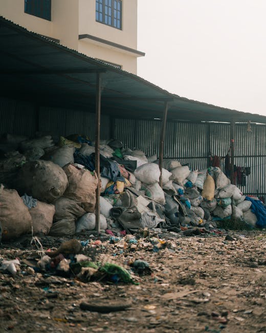 An expansive landfill site with large piles of mixed waste material, including plastic bags, paper, and other debris, piled high on a sloping hill. Multiple yellow excavators are actively engaged in m